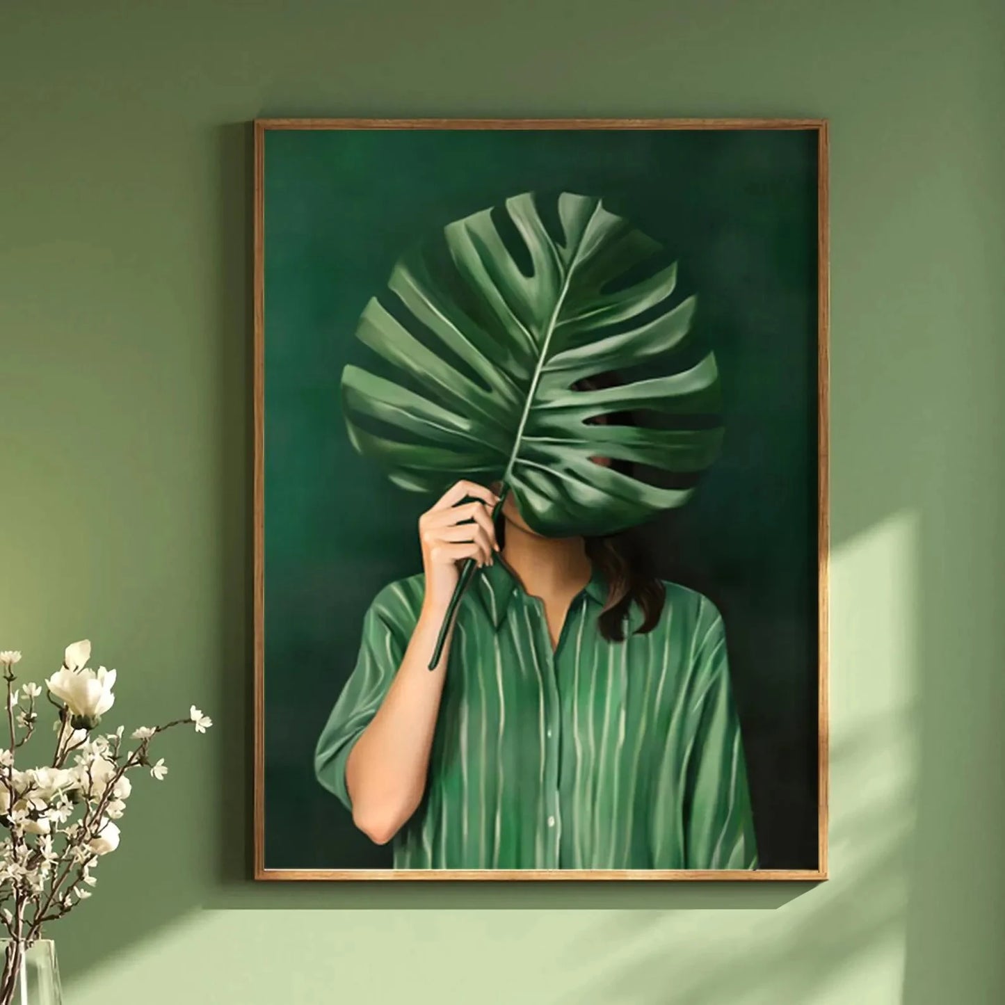 Framed artwork of a person holding a large green leaf over their face against a green wall.