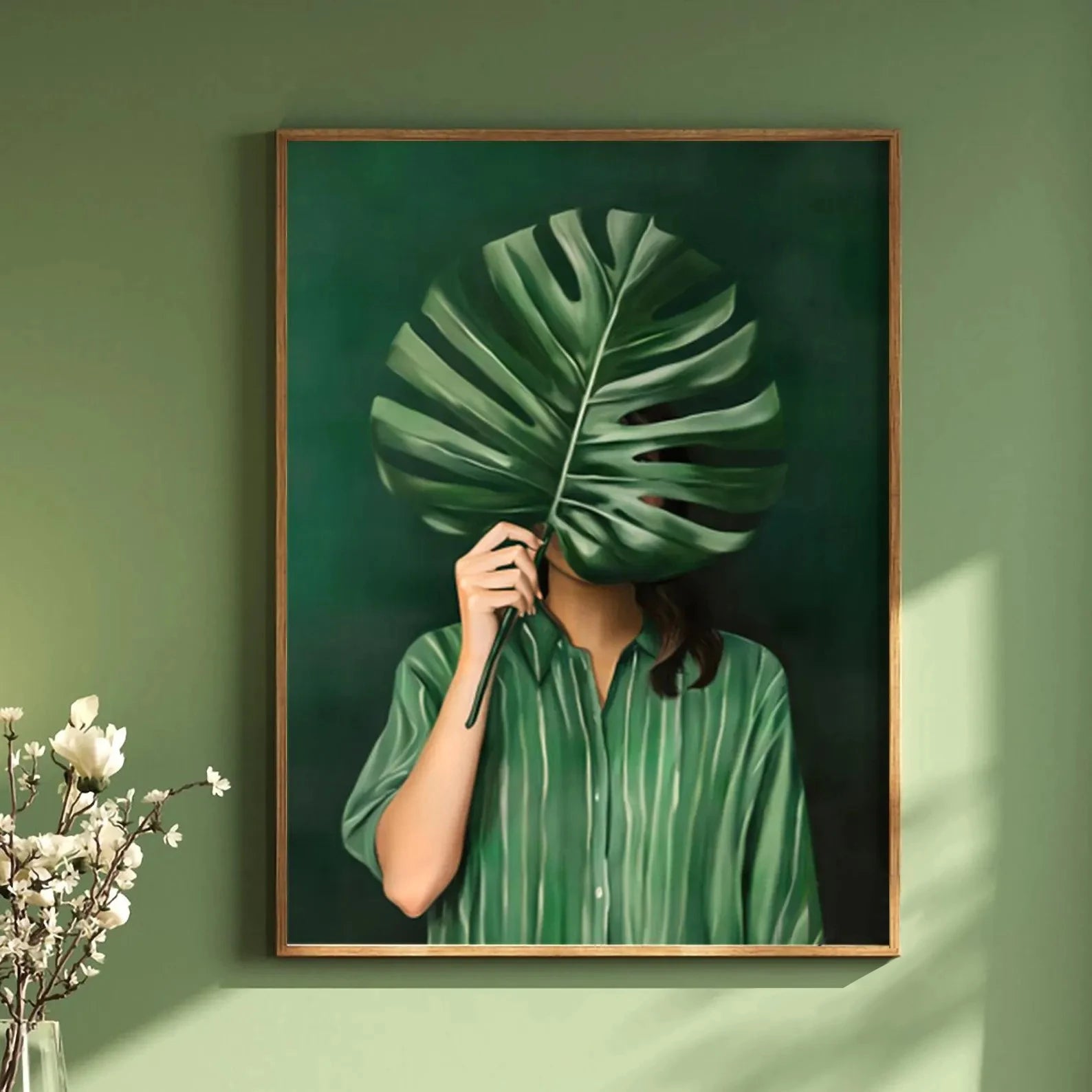 Framed artwork of a person holding a large green leaf over their face against a green wall.