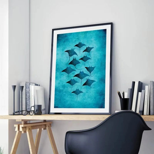 Framed artwork of manta rays on a teal background above a desk with books and a chair. Canvas & Curio