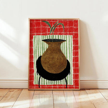 Framed artwork of a vase with a plant on a red and green striped background, leaning against a white wall.