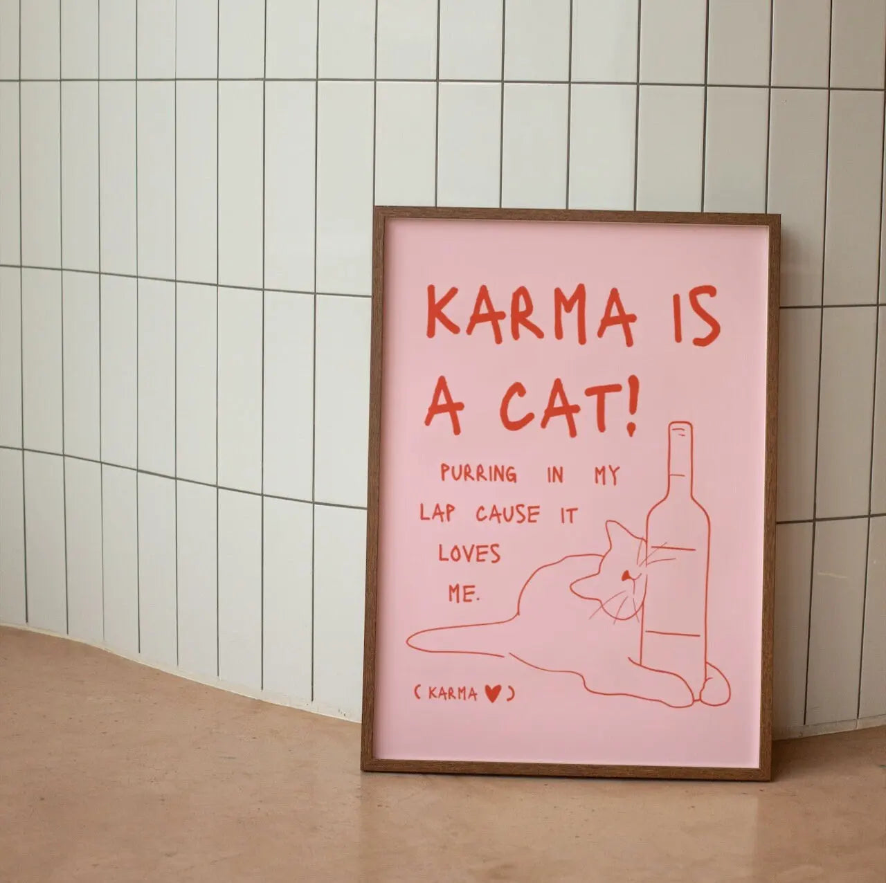 Framed pink poster with text 'Karma is a cat! Purring in my lap cause it loves me.' against a tiled wall.
