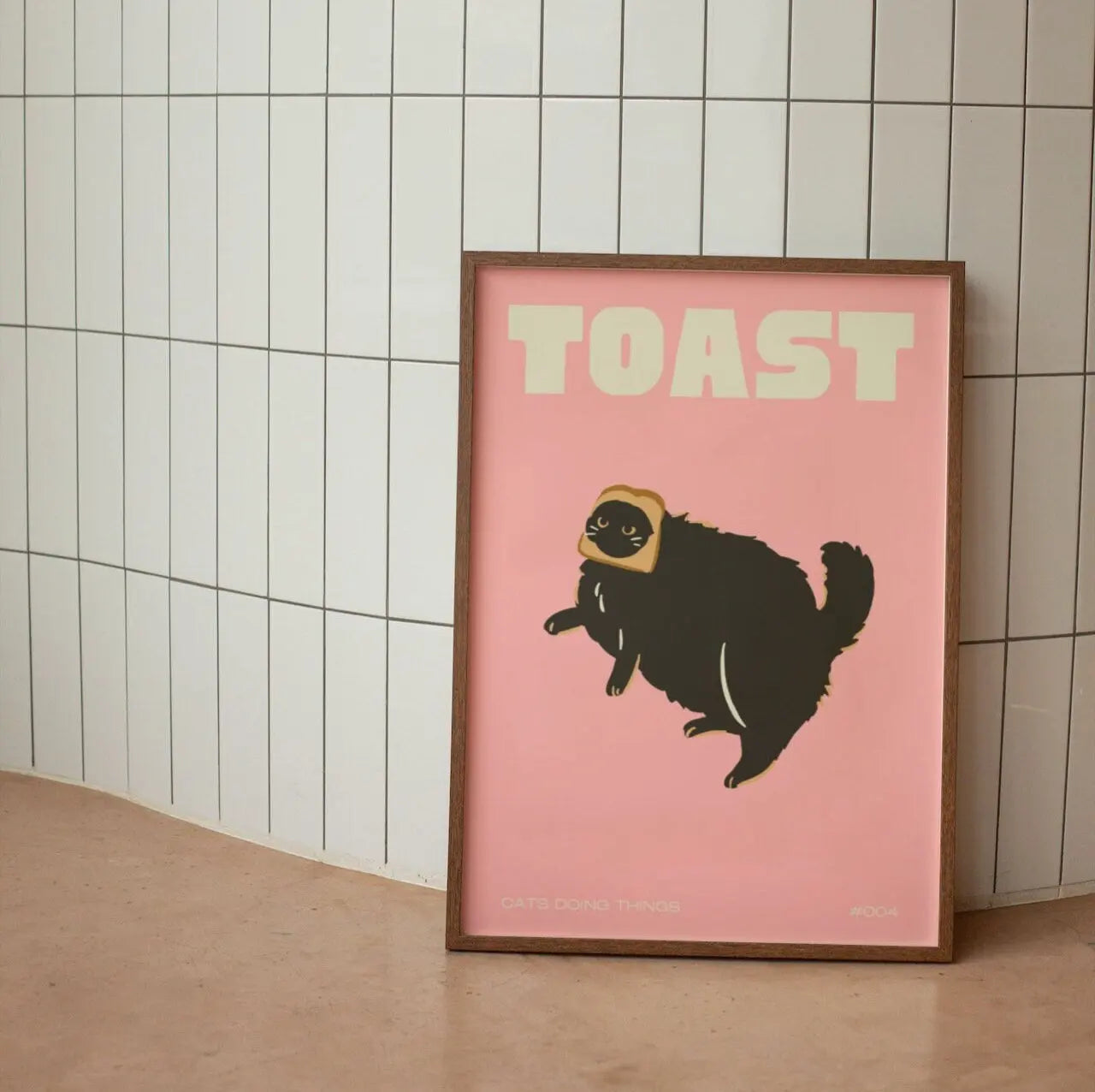 Framed artwork with a black cat illustration and 'TOAST' text on a pink background, leaning against a tiled wall.