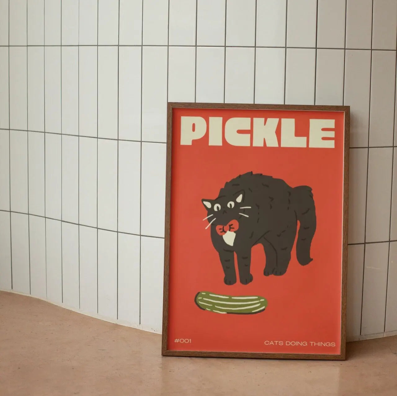 Framed poster with a black cat and pickle illustration on an orange background, leaning against a tiled wall.