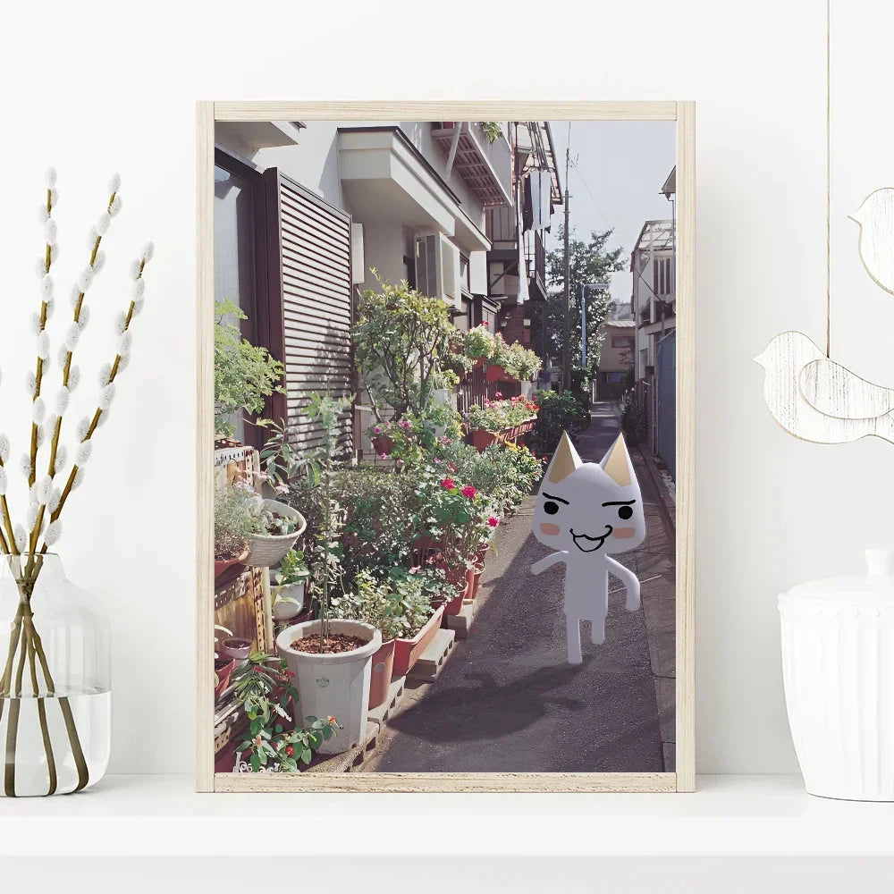 Framed picture of a narrow street with plants and a cartoon cat character on a white wall.