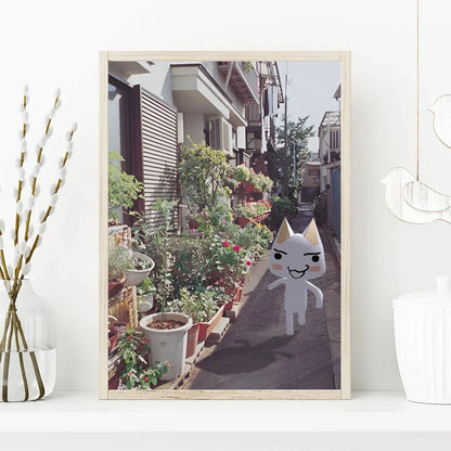 Framed picture of a narrow street with plants and a cartoon cat character on a white wall.